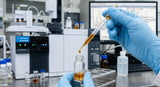 A close-up, high-definition photograph of a technician's gloved hand holding a glass pipette, carefully dropping a single drop of rich, amber-gold Jasmine Grandiflorum Absolute into a clear glass vial for GC-MS analysis. The background shows blurred but sophisticated laboratory equipment, including a gas chromatograph machine and computer screens displaying complex chemical charts. The lighting is precise and clean, emphasizing the purity and concentration of the oil.