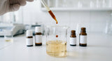 A close-up shot on a clean, laboratory formulation bench. A clear glass beaker holds a pale golden facial oil. A glass pipette is actively dispensing a drop of dark amber frankincense essential oil into the beaker, creating subtle ripples. In the softly blurred background, there are small, amber glass cosmetic bottles with blank labels.