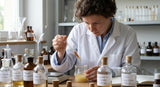 A close-up laboratory scene showing a perfumer in a white coat carefully adding a drop of viscous, amber-colored Kewra absolute from a glass pipette into a complex mixture in a beaker, surrounding glass vials and equipment, focused expression, soft background blur