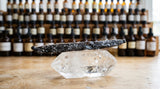 A macro photograph focusing on a single, dark, crystallized vanilla bean resting on a piece of clear quartz. In the background, a professional perfumer's organ with hundreds of small bottles is visible but softly blurred. The lighting is natural daylight, highlighting the texture of the bean and the clarity of the quartz.