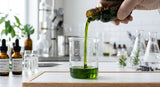 A sophisticated, close-up photograph capturing the viscosity and rich, natural color of premium cold-pressed avocado oil being gently poured into a clear glass beaker. The background is a clean, modern cosmetic laboratory setting with stainless steel equipment and blurred botanical elements, suggesting purity and precision manufacturing.