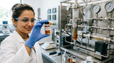 A close-up shot of a scientist in a clean laboratory setting, wearing gloves and safety glasses, holding up a glass vial containing a rich, amber-colored Lavender Co2 Extract oil. The background is a slightly blurred stainless steel supercritical extraction unit.