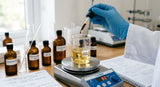 A close-up photograph of a chemist's gloved hand carefully adding a single drop of dark amber bakuchi oil from a glass pipette into a beaker containing a pale gold carrier oil blend. The beaker sits on a magnetic stirrer hotplate, which is gently mixing the liquids. In the background, out of focus, are various other cosmetic raw materials in amber bottles and a lab notebook with handwritten formulations. The lighting is bright and clean, typical of a professional formulation lab, emphasizing precision.