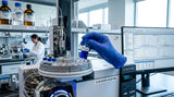 Laboratory setting. A close-up shot of a gas chromatograph (GC-MS) machine in operation. A gloved hand inserts a vial containing a deep blue liquid. The background is a clean, modern lab with equipment and a computer screen showing complex data graphs. The lighting is bright and clinical.