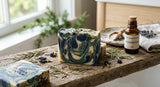 A close-up photograph of artisanal cold process soap bars with a marbled pattern, resting on a rustic wooden plank, with scattered dried cedar needles and a small bottle of oil in the background