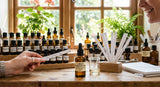 Close-up photograph of a perfumer's organ, with a bottle labeled 'Linden Blossom Abs. (Low Eugenol)' prominent in the foreground, surrounded by other vials, a glass beaker, and professional smelling strips, backlit by soft, natural light coming through a window