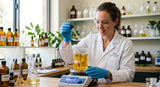 A laboratory setting with a researcher in a white coat and gloves carefully adding a clear, pale yellow liquid from a glass dropper into a beaker containing a rich, golden-colored oil. The beaker is on a magnetic stirrer, and the liquids are swirling together. Various other glass beakers and flasks containing different botanical extracts and oils are visible in the background, out of focus.