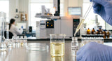 A macro photograph capturing a glass beaker containing a clear, pale yellow liquid, with a small amount being carefully pipetted into a GC-MS vial. The background is a clean, well-lit laboratory environment with analytical equipment and glassware, all slightly out of focus. A subtle glow emanates from the instruments, suggesting activity and precision.