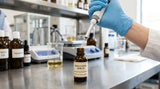 A close-up photograph of a gloved hand using a micro-pipette to carefully dispense a pale amber liquid into a small, dark glass dropper bottle. The bottle is labeled 'Bakuchiol Serum'. The setting is a clean, well-organized cosmetic formulation lab, with stainless steel equipment and glass beakers partially visible in the background. The lighting is focused and professional.