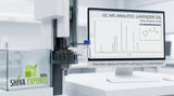 A close-up of a modern gas chromatography mass spectrometer in a clinical, white-lit laboratory, showing a glass autosampler vial containing a clear liquid, with a computer monitor displaying a sharp chromatogram peak in the background