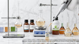 A clinical laboratory glassware setup with a pale amber synthetic aroma chemical liquid being pipetted into a clear glass vial, white marble background, scientific aesthetic