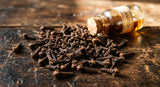 A macro photograph capturing the rough texture of dried clove buds spilled onto a dark, aged wooden surface. In the background, a small glass vial with a cork stopper is partially visible, bathed in warm, directional light that highlights the rich browns and hints of amber.