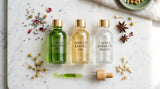 A serene, overhead photograph on a polished light marble surface, displaying three distinct clear glass vials. One vial contains rich green cold-pressed avocado oil, another a pale gold apricot kernel oil, and the third a clear essential oil blend. They are arranged neatly with a minimalist pipette and a few scattered dried botanicals, representing premium cosmetic raw materials. The lighting is soft and natural, emphasizing the textures and colors.