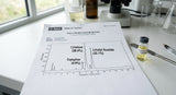 A close-up photograph of a gas chromatograph printout showing distinct peaks for linalool, linalyl acetate, and camphor, lying on a clean white laboratory bench next to a small glass vial filled with pale yellow lavandin oil. Natural light illuminates the scene.