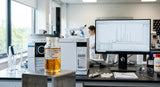 A close-up photograph of a professional laboratory setting, focusing on a clear glass vial containing a rich, amber-colored Jasmine Sambac Absolute. The vial is positioned next to a modern GC-MS machine with a computer monitor displaying complex chromatogram peaks in the background. Natural light from a window illuminates the scene, highlighting the viscosity of the absolute.