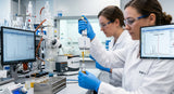 A close-up, sterile laboratory shot of a scientist in a white lab coat using a micropipette to carefully drop a clear, golden liquid into a glass vial. The background shows complex glassware, a rotary evaporator, and a computer screen displaying a chromatogram with a prominent peak. The lighting is bright and clinical, emphasizing precision and purity.