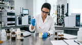 A sophisticated laboratory setting with a stainless steel bench. A formulator in a white coat and nitrile gloves is using a precision glass pipette to add a drop of viscous, amber-colored Jasmine Grandiflorum Absolute into a beaker containing a cream base. The background shows complex distillation glassware and analytical equipment, suggesting a focus on QC and precise formulation.