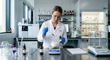A sterile laboratory setting with a stainless steel workbench. A formulator, wearing gloves and a lab coat, is carefully using a glass pipette to add a drop of oil from a brown bottle labeled 'Bergamot FCF' into a beaker containing a cream base. Various analytical instruments and beakers are visible in the background, suggesting a precise and controlled formulation process.