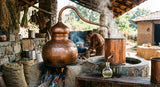 A close-up photograph of a traditional copper pot still used for essential oil distillation in rural India, with steam rising and condensed oil dripping into a glass receiver, set in a rustic, open-air distillery environment.