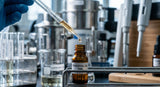 A macro photograph focusing on the tip of a glass perfumer's pipette dispensing a single, clear drop of liquid into a small, amber glass vial. The background is a gently blurred laboratory bench with stainless steel equipment, bathed in cool, analytical light.