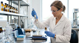 A detailed, high-resolution photograph of a cosmetic scientist in a clean, modern laboratory, wearing a white lab coat and gloves, using a micropipette to carefully add a pale yellow oil from a glass vial into a small beaker containing a clear serum base. The background is slightly blurred with stainless steel equipment and glass beakers, focused on the precise action.