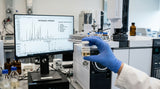 A close-up photograph taken in a modern fragrance laboratory. A gloved hand holds a small, clear glass vial containing a pale, amber-colored liquid, labeled 'Ambergris Oil'. The vial is positioned in front of a complex Gas Chromatography-Mass Spectrometry (GC-MS) machine, with a computer screen in the background displaying a detailed chromatogram chart with multiple peaks, indicating the chemical analysis of the sample. The lighting is sterile and precise.