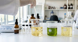 A close-up, high-definition photograph of a lab bench with several glass beakers. One beaker contains a clear, pale golden oil (bakuchiol) being stirred with a glass rod. Another beaker holds a vibrant green oil (cucumber seed oil). A third beaker contains a pale yellow oil (evening primrose oil). The scene is bathed in bright, clean, diffused light, highlighting the textures and colors of the oils and the clarity of the glassware.