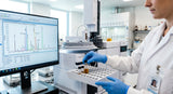 A close-up photograph of a lab technician in a white coat and blue nitrile gloves holding a clear glass vial containing a golden-brown oil. The technician is placing the vial into a sample tray of a modern gas chromatography-mass spectrometry (GC-MS) instrument. In the background, a computer monitor displays a complex chromatogram with multiple peaks. The lighting is bright and clean, typical of a professional laboratory.