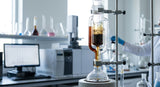 A close-up laboratory photograph showing a glass Soxhlet extraction apparatus in operation. A rich, dark brown vanilla extract is cycling through the glass chamber, illuminated by a soft, clean white light. In the background, out of focus, are various volumetric flasks and a GC-MS instrument console. The atmosphere is precise and scientific.