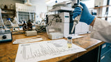 A close-up view of a GC-MS chromatogram printout lying on a lab bench. A gloved hand holds a micropipette over a small vial containing a pale yellow liquid, presumably rose oil. In the background, out of focus, is a complex gas chromatograph instrument. The overall impression is one of rigorous, scientific quality control.