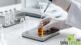 A close-up shot of a researcher using a precise micropipette to drop a clear liquid into a small glass vial containing amber-colored essential oil, clean white laboratory bench, stainless steel instruments in background