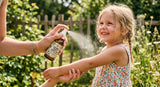 A close-up shot of a hand applying a natural insect repellent spray (clearly labeled with clove oil) to a child's arm outdoors, with focus on the spray mist and the child's happy expression