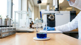 A close-up photograph of a clear glass beaker containing a small amount of deep blue essential oil, with a dropper suspended just above it. The background is a clean, modern laboratory setting, with stainless steel equipment and a blurred GC-MS machine in the distance. The lighting is bright and clean.