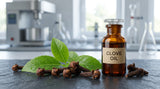 Macro photograph of dried clove buds and a few fresh green leaves on a dark, textured slate surface. A small, amber glass vial with a glass stopper sits beside them, catching the light. The background is a gently blurred laboratory setting with stainless steel equipment and glass beakers, illuminated by soft, natural daylight from a window, creating a precise and clinical atmosphere.