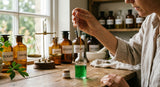 A close-up photograph of a perfumer's hand using a glass pipette to carefully add a drop of viscous, amber-colored Galbanum resin into a clear glass vial containing a vibrant green liquid. The background is softly blurred, showing other apothecary bottles and a vintage brass scale, bathed in warm, diffused light from a nearby window.