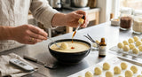 A close-up, high-angle photograph of professional-grade white chocolate truffles being dipped into a tempering bowl. A pipette is adding a drop of rich amber vanilla oil into the melted white chocolate pool. The setting is a clean, modern chocolatier's kitchen with stainless steel surfaces and diffused light.