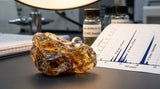 A detailed macro photograph capturing the texture of a rugged, raw Galbanum resin tear, showcasing its internal, translucent, golden-brown layers. A single, perfect drop of water rests on its surface, reflecting the soft, warm light of a laboratory setting. In the background, a portion of a GC-MS printout with sharp, defined peaks is visible but softly out of focus, emphasizing the analytical verification of the material's purity.
