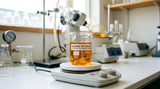 A macro photograph focused on a clear glass beaker containing a rich, amber-colored liquid. The beaker sits on a white laboratory workbench. In the background, out of focus, are various scientific instruments like a magnetic stirrer and a rotary evaporator. The light is bright and clean, emphasizing the clarity and color of the Bakuchiol 99% isolate.