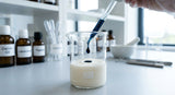 A close-up studio photograph of a laboratory beaker containing a pale, unscented cosmetic cream base. A glass dropper is suspending a single, perfect drop of deep, inky blue German chamomile oil just above the cream's surface, preparing to mix. The background is a clean, modern lab bench with other formulation tools, gently blurred. The lighting is crisp and cool, emphasizing the contrast between the white cream and the intense blue oil.